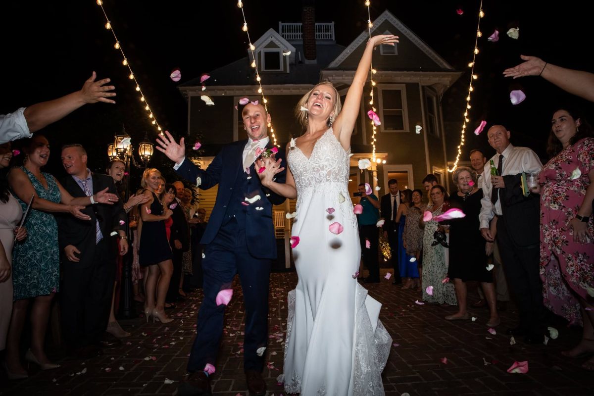 A bride and groom laughing through a shower of rose petals under string lights as guests cheer