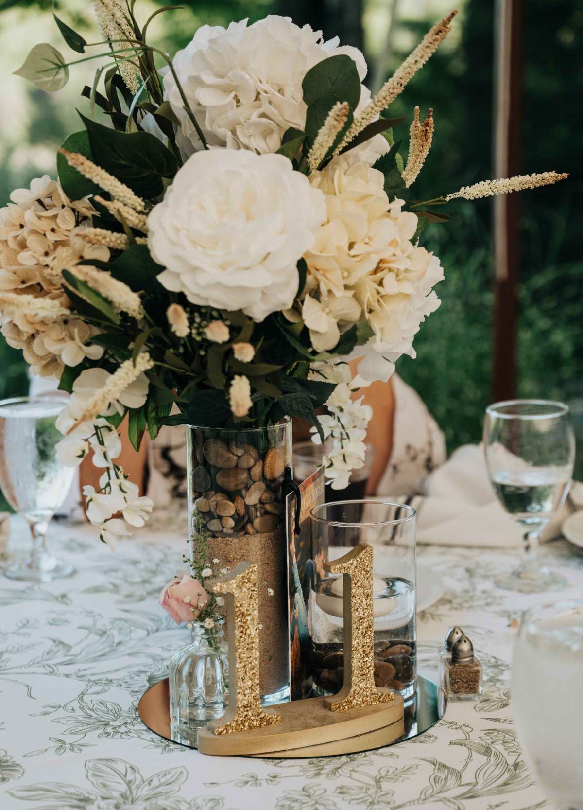 A wedding table centerpiece of white peonies and hydrangeas in a stone-filled vase with a gold glitter table number 11